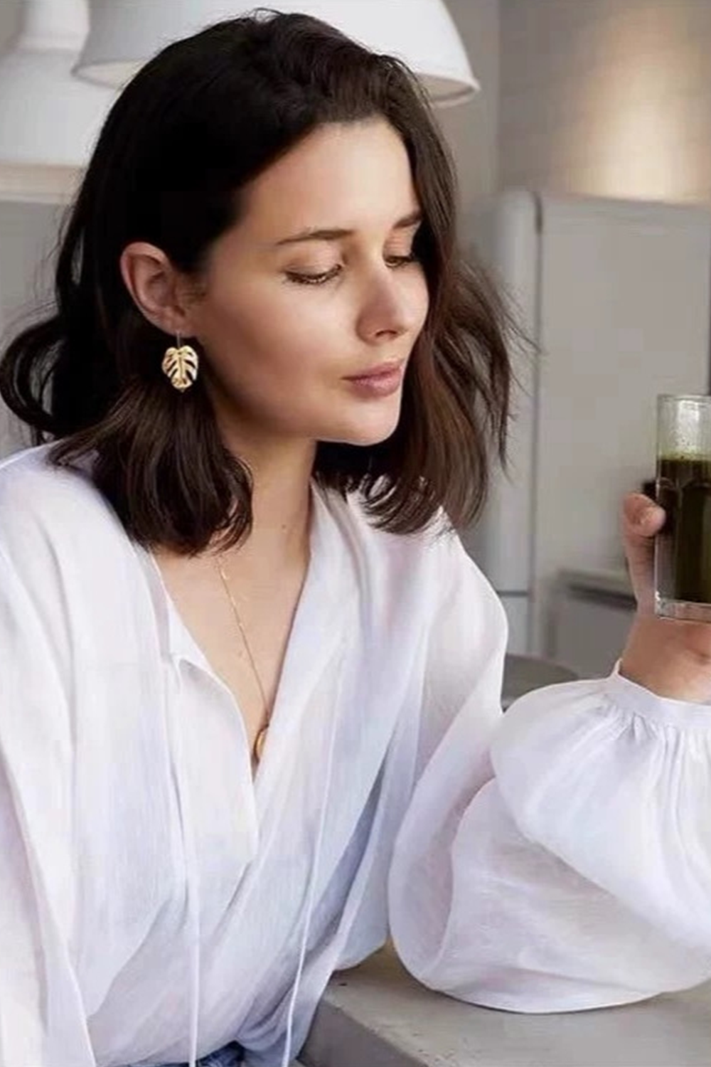 Brunette in white top wearing monstera hoops, holding matcha latte - Tropical-luxe home aesthetic​